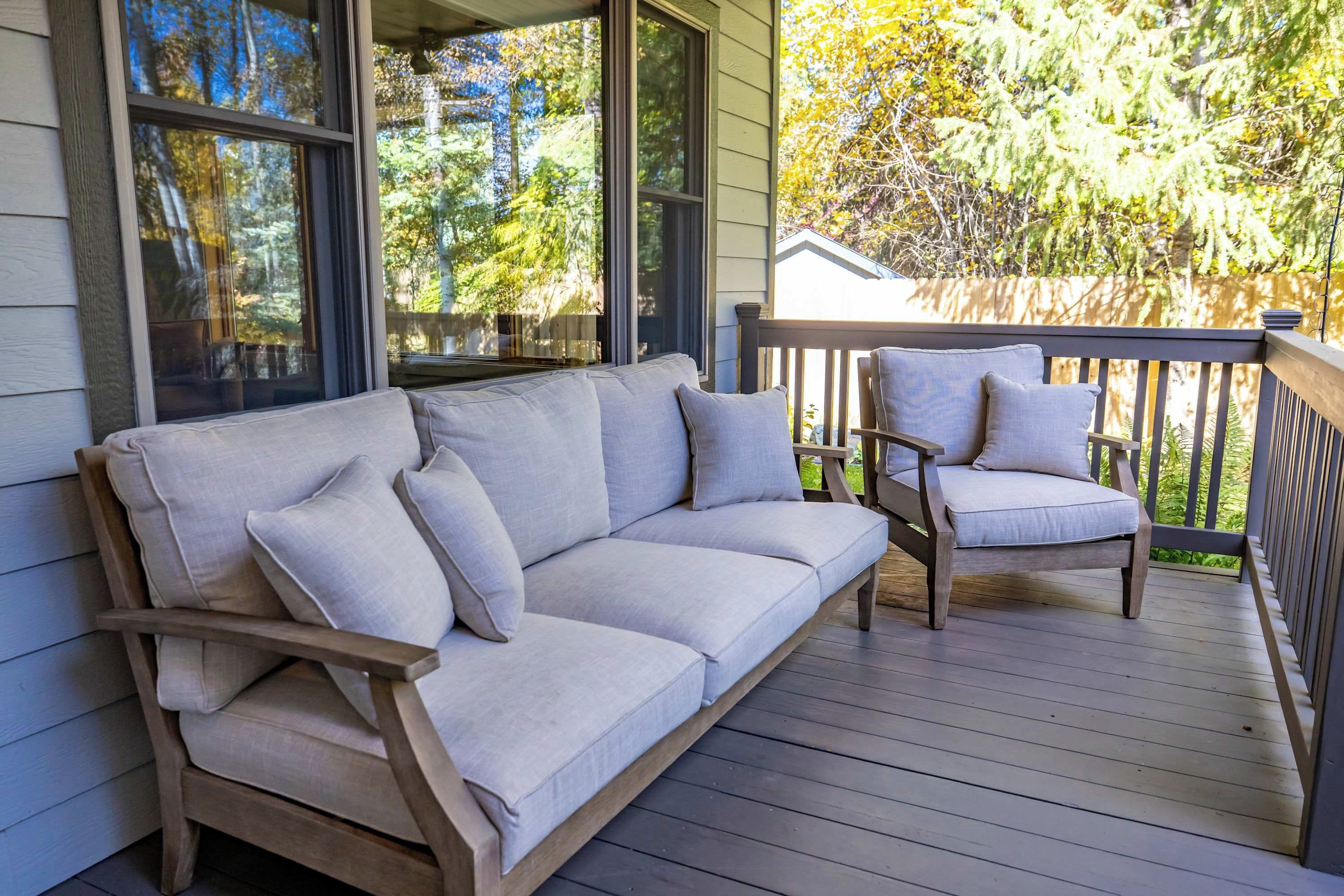 Eine Holzterrasse mit einer Couch und zwei Stühlen, einem Geländer aus Holz und Blick auf Bäume sowie ein Haus.