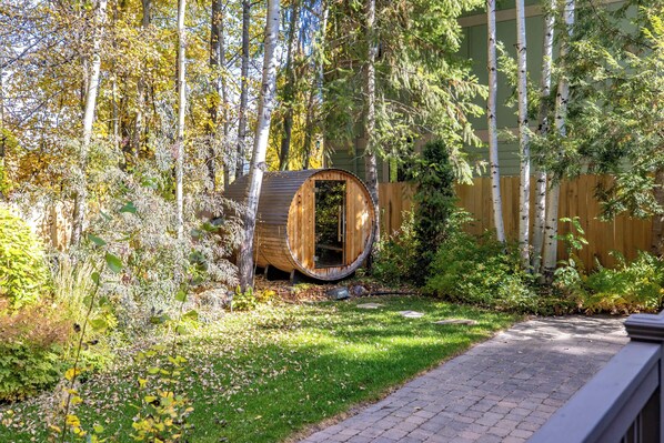 A wooden barrel structure in a garden with a wooden fence and trees in the background. - Seasonal Retreat | Private Yard w/Sauna & Spa (Whitefish)