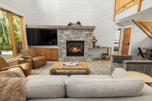 A modern living room with a stone fireplace, comfortable seating, a flat-screen TV, and a view of the outdoors. - Seasonal Retreat | Private Yard w/Sauna & Spa (Whitefish)