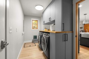 Cocina funcional con armarios grises, encimera de madera, lavadora y ventana con vista a árboles.