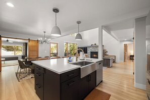 A modern kitchen with a large island, pendant lighting, and a stone fireplace. - Seasonal Retreat | Private Yard w/Sauna & Spa (Whitefish)