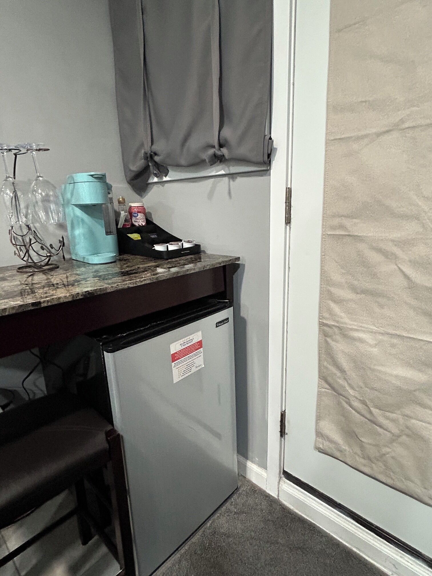 A compact kitchenette with a refrigerator, a small sink, and a countertop. There are wine glasses and a blue water bottle on the counter.