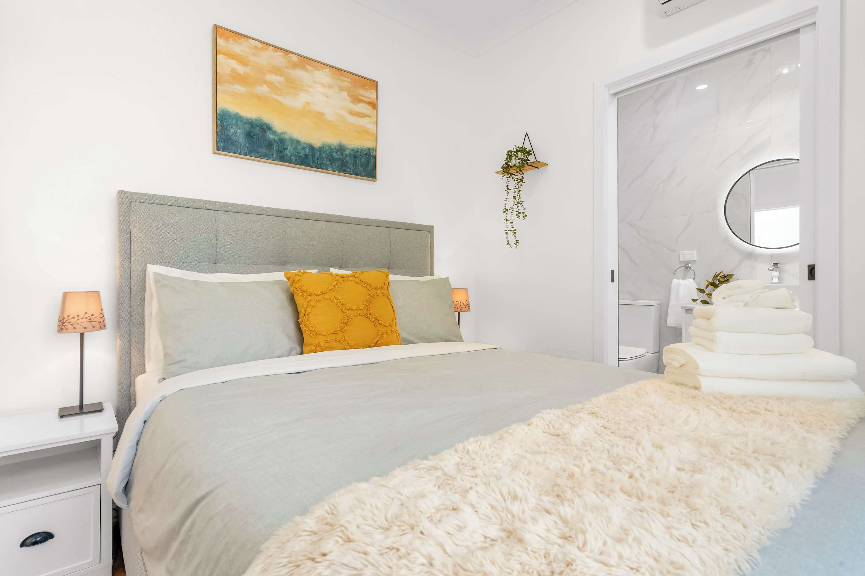 A modern bedroom with a bed, bedside lamps, a fluffy rug, a wall-mounted plant, and a bathroom with a mirror and towel rack.