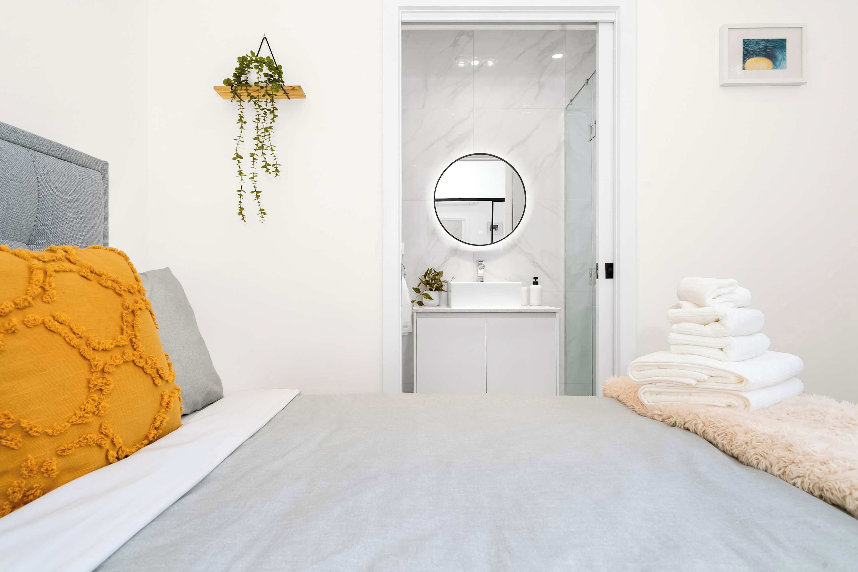 Quarto com cama, mesa de cabeceira, planta pendurada, banheiro com espelho e uma pilha de toalhas.