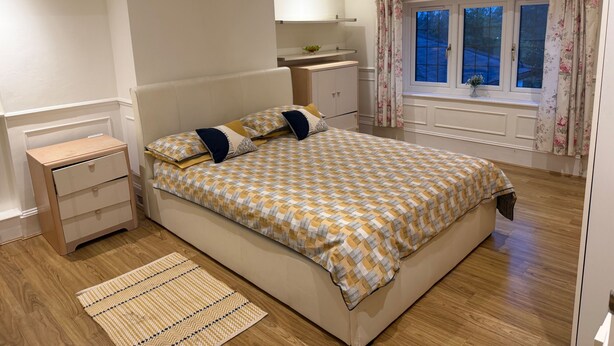 A bedroom with a bed, bedside tables, a nightstand, a window with curtains, and a small table with a plant.