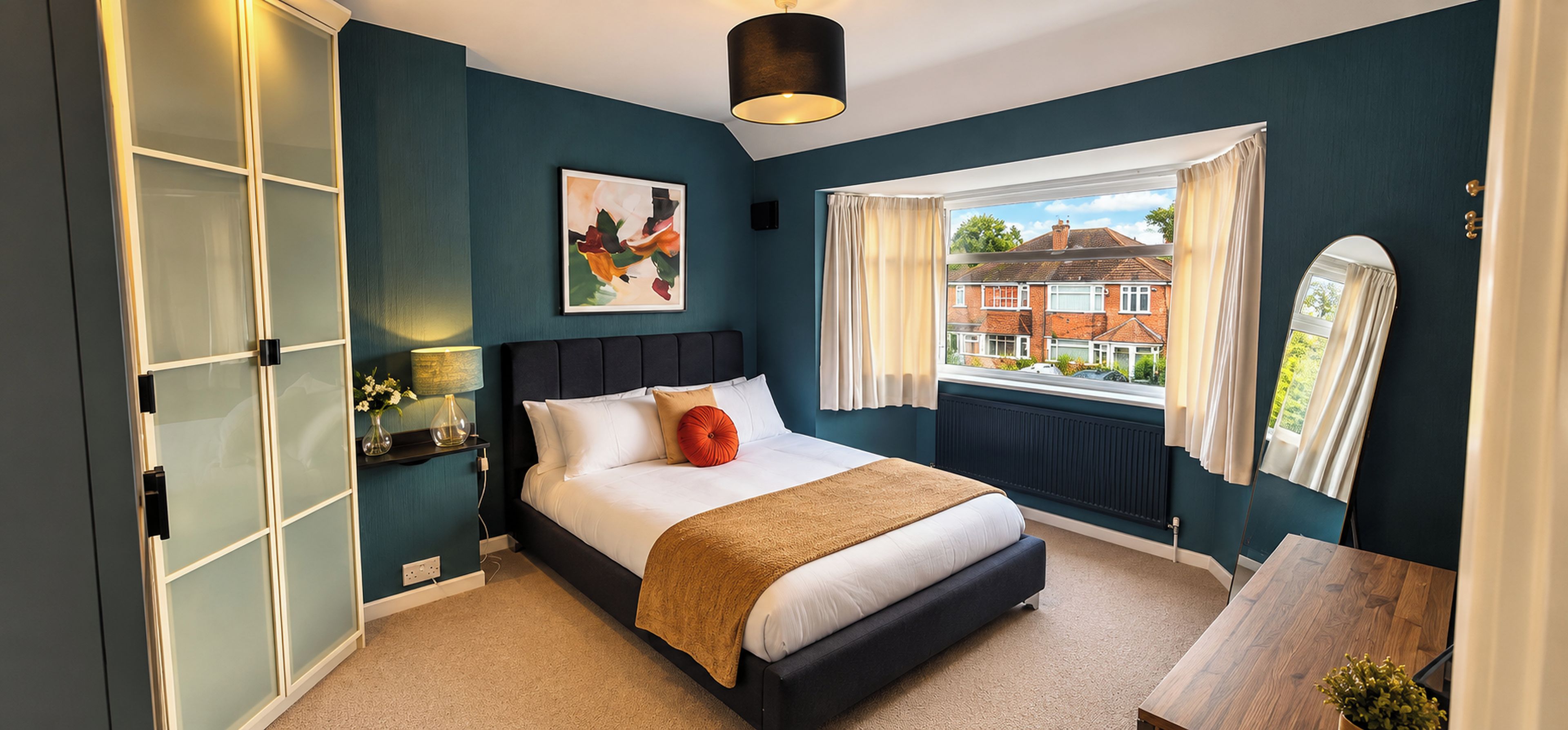 A bedroom with a bed, a window with curtains, a radiator, and a view of houses outside.