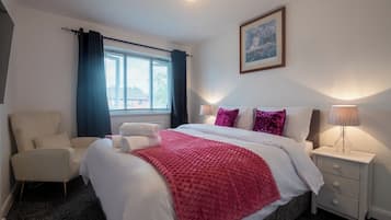 A hotel room with a bed, a chair, a bedside table with a lamp, a window with curtains, and a framed picture on the wall.