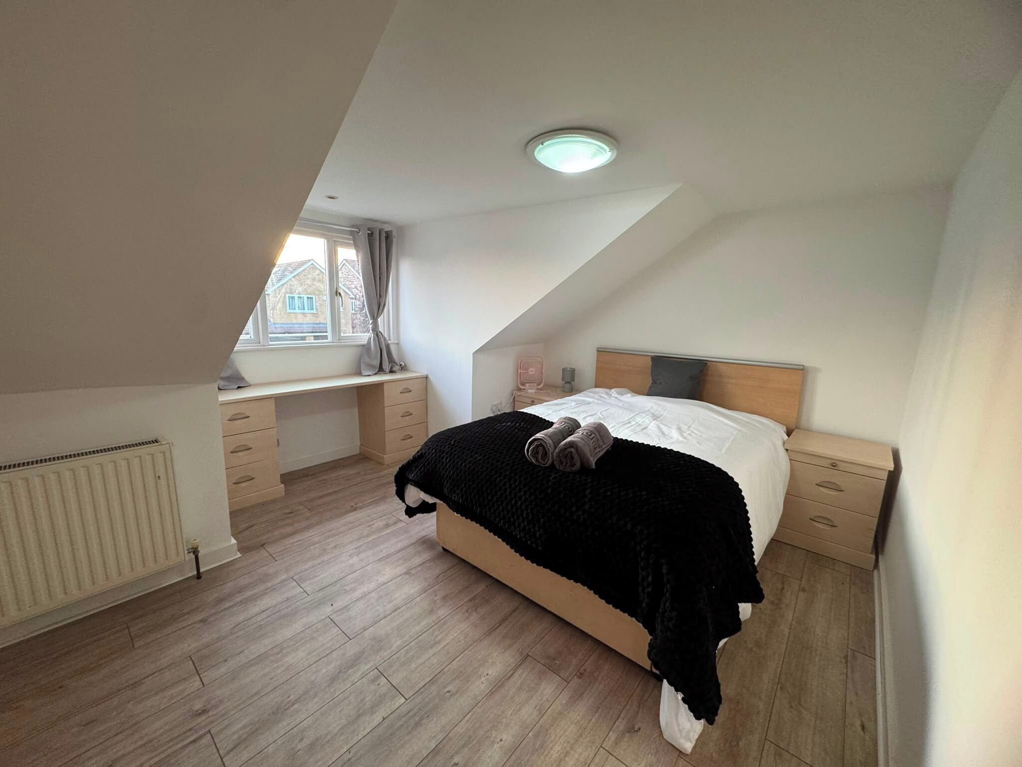 Une chambre avec un plafond en pente, un lit recouvert d’une couverture noire, une table de chevet avec une lampe et une fenêtre avec des rideaux.