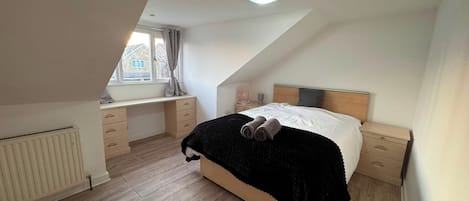 A bedroom with a sloped ceiling, a bed with a black blanket, a bedside table with a lamp, and a window with curtains.