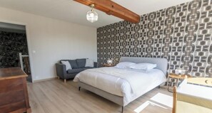 A bedroom with a bed, a grey sofa, a wooden headboard, a patterned wall, and a wooden floor.
