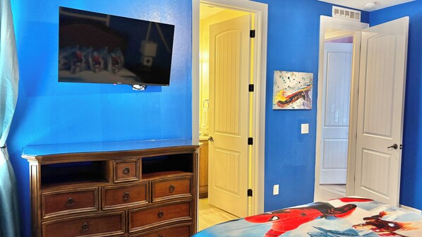Ein Zimmer mit blauen Wänden, einem Holzschrank, einem Bett mit Spider-Man Bettwäsche, einem an der Wand befestigten Fernseher und einer Tür zu einem anderen Raum.