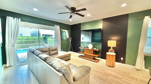A modern living room with a sofa, a coffee table, and a TV. There is a ceiling fan and a window with curtains. - Spacious 8BR Disney-Area Home| Pool & Home Theater (Kissimmee)