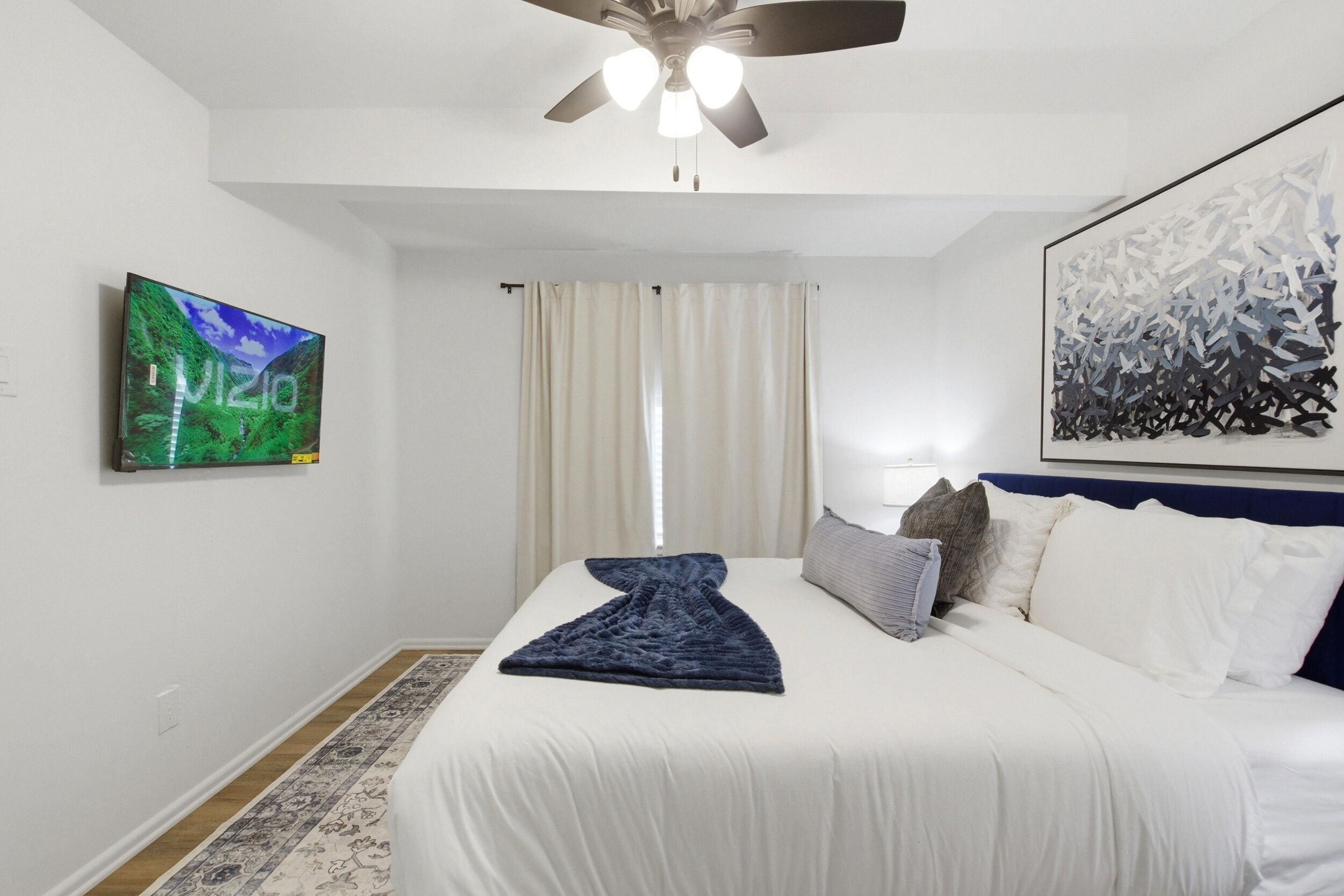 Una habitación de hotel con una cama, un ventilador de techo, un televisor en la pared y un cuadro abstracto grande.