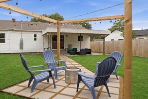 Eine Hinterhofterrasse mit Adirondack-Stühlen, einem Metallgrill und einer Pergola mit Lichterketten.