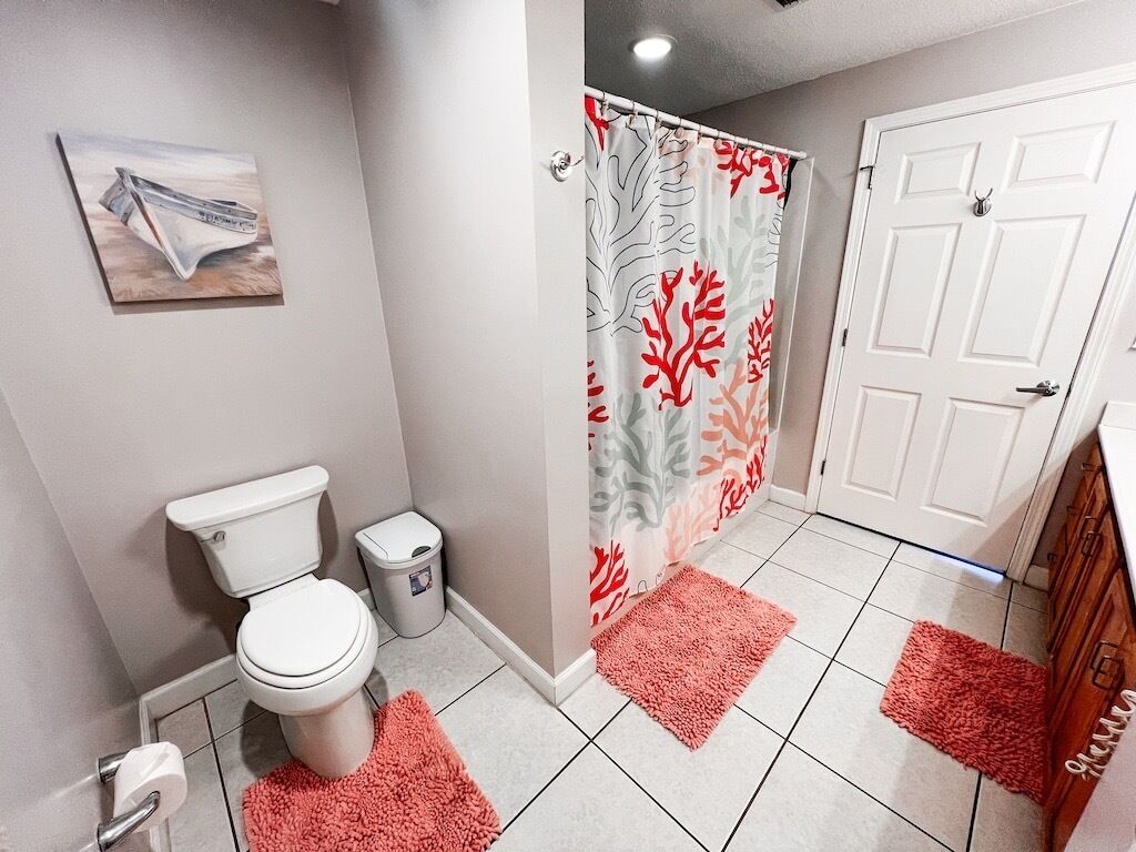 Un baño con inodoro, un cubo de basura, una cortina de ducha con estampado de coral y una alfombra rosa.