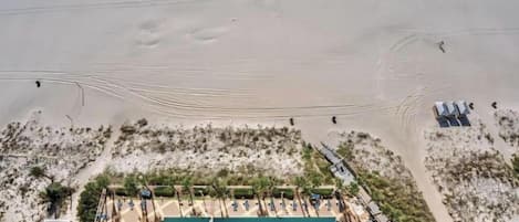 Vista aérea de un complejo turístico en la playa con piscina, sombrillas y algunas personas caminando sobre la arena.