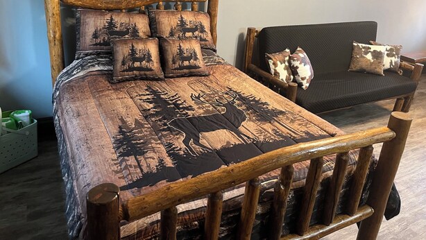 A wooden bed with a forest-themed bedspread and matching pillows, a black sofa with a cow print pillow, and a wooden chair with a woven seat.