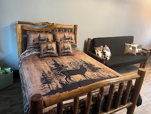 A wooden bed with a forest-themed bedspread and matching pillows, a black sofa with a cow print pillow, and a wooden chair with a woven seat.