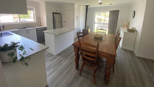 A modern kitchen with white cabinetry, a dining area with a wooden table and chairs, and a view of the outdoors through a window. -     Your Home Away From Home (Inverell)