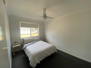 Un dormitorio con una cama, ventilador de techo, ventana y una mesita de noche.