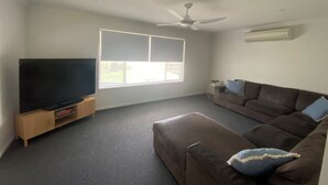 A living room with a flat-screen TV on a wooden stand, a brown leather sofa with blue cushions, a ceiling fan, and a large window with blinds. -     Your Home Away From Home (Inverell)