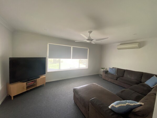 A living room with a flat-screen TV on a wooden stand, a brown leather sofa with blue cushions, a ceiling fan, and a large window with blinds.