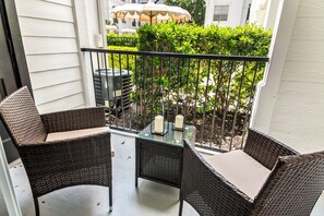 A balcony with wicker furniture and a glass table, overlooking a garden with a grill and umbrellas.