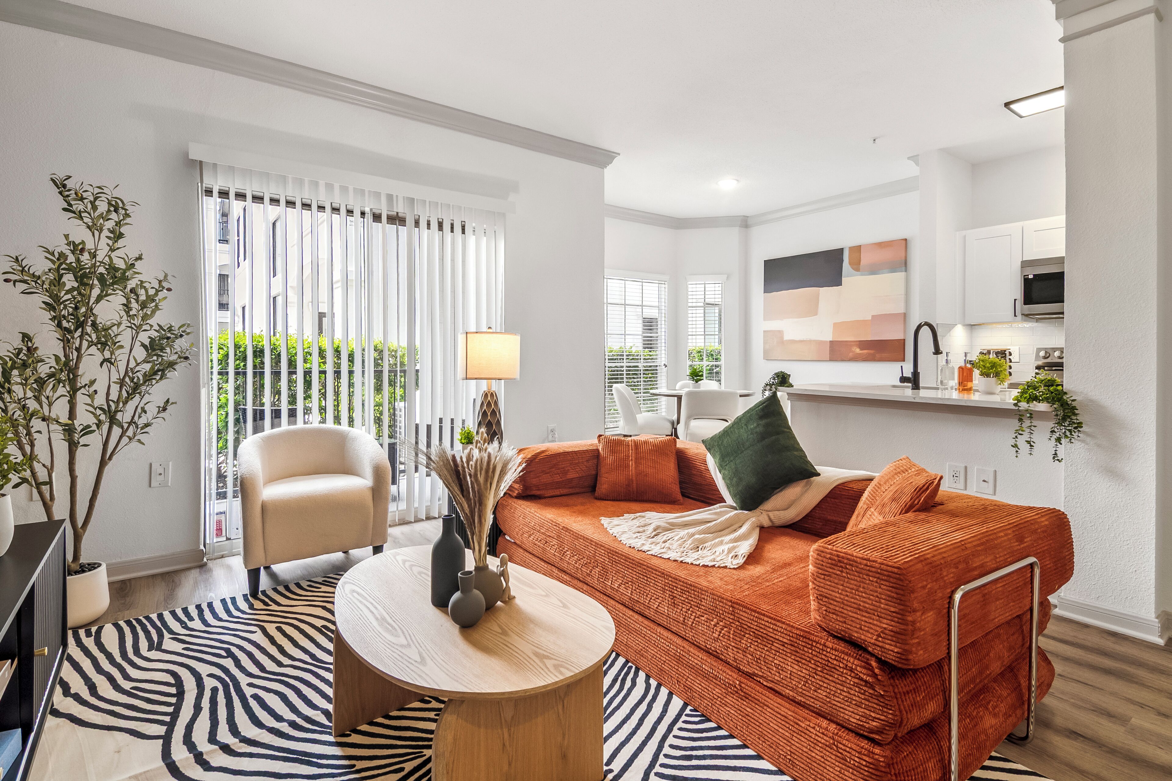Un salon moderne avec un canapé, un fauteuil et une table basse ronde. Une grande fenêtre avec des stores, une plante en pot et un coin cuisine se trouvent à l’arrière-plan.