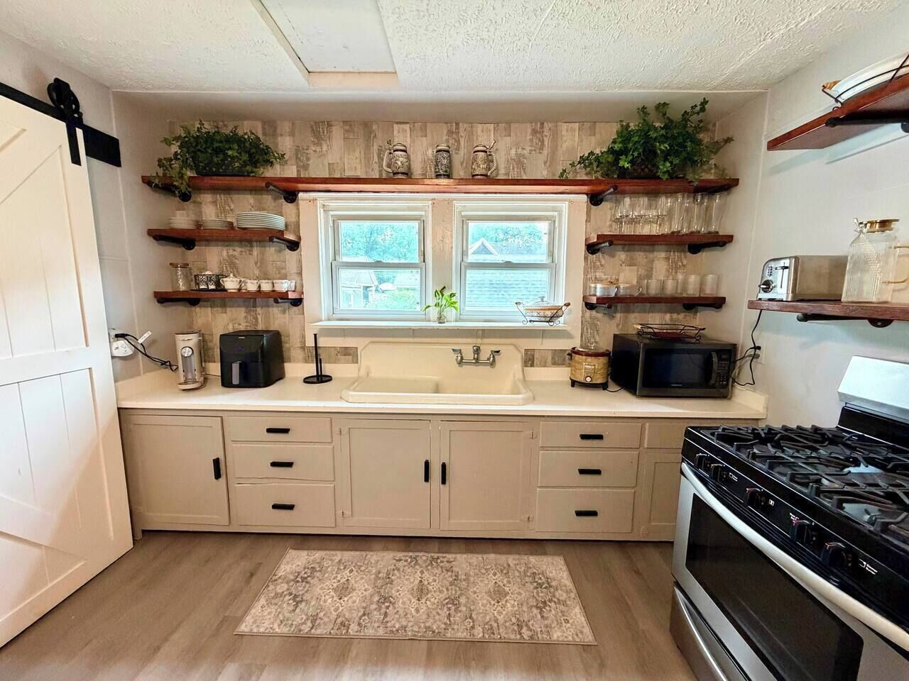 A kitchen with a double sink, wooden cabinets, a stove, and shelves with plants and kitchenware.