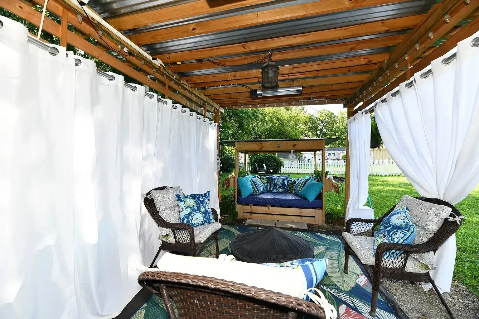 A covered patio with a bed, two wicker chairs, and a table.