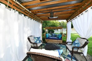 A covered patio with a bed, two wicker chairs, and a table.