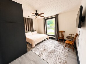 A bedroom with a bed, a ceiling fan, a window with curtains, a small table with a candelabra, and a chair.