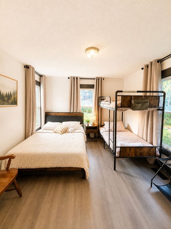 A room with a single bed, a bunk bed, a wooden floor, a window with curtains, and a painting on the wall.