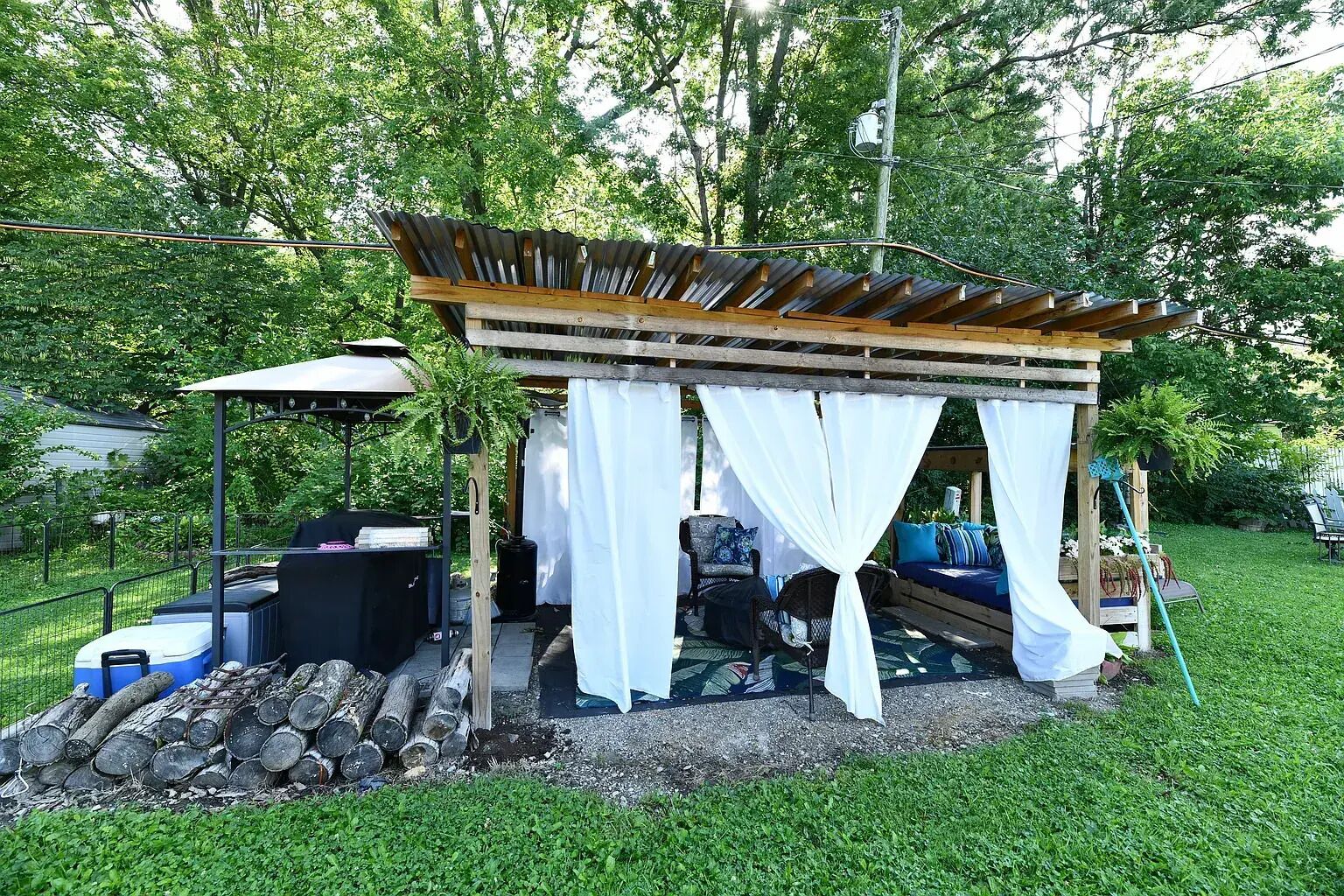 A wooden gazebo with a covered seating area, a grill, and a pile of firewood.