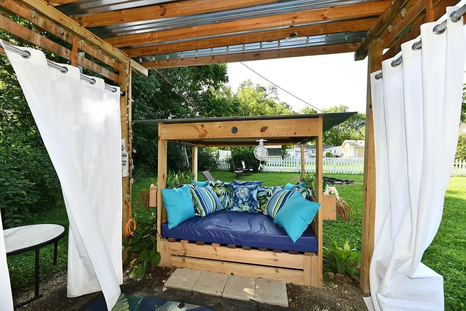 A wooden swing bed with cushions, a fan, and a white fence in the background.