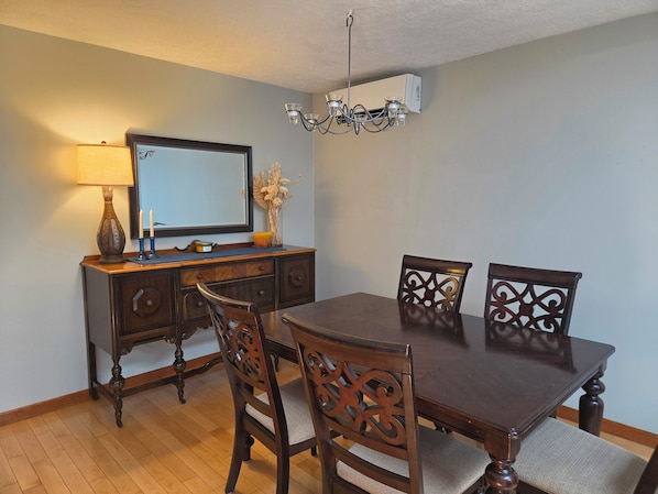 A dining area with a wooden table, four chairs, a television on a wooden cabinet, a chandelier, and a decorative vase with dried flowers.