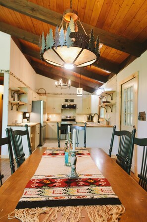 A dining area with a long wooden table, chairs, a chandelier, and a kitchen in the background.