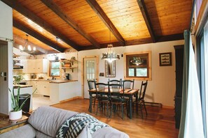A spacious living area with a wooden ceiling, a dining table with chairs, and a kitchen in the background.