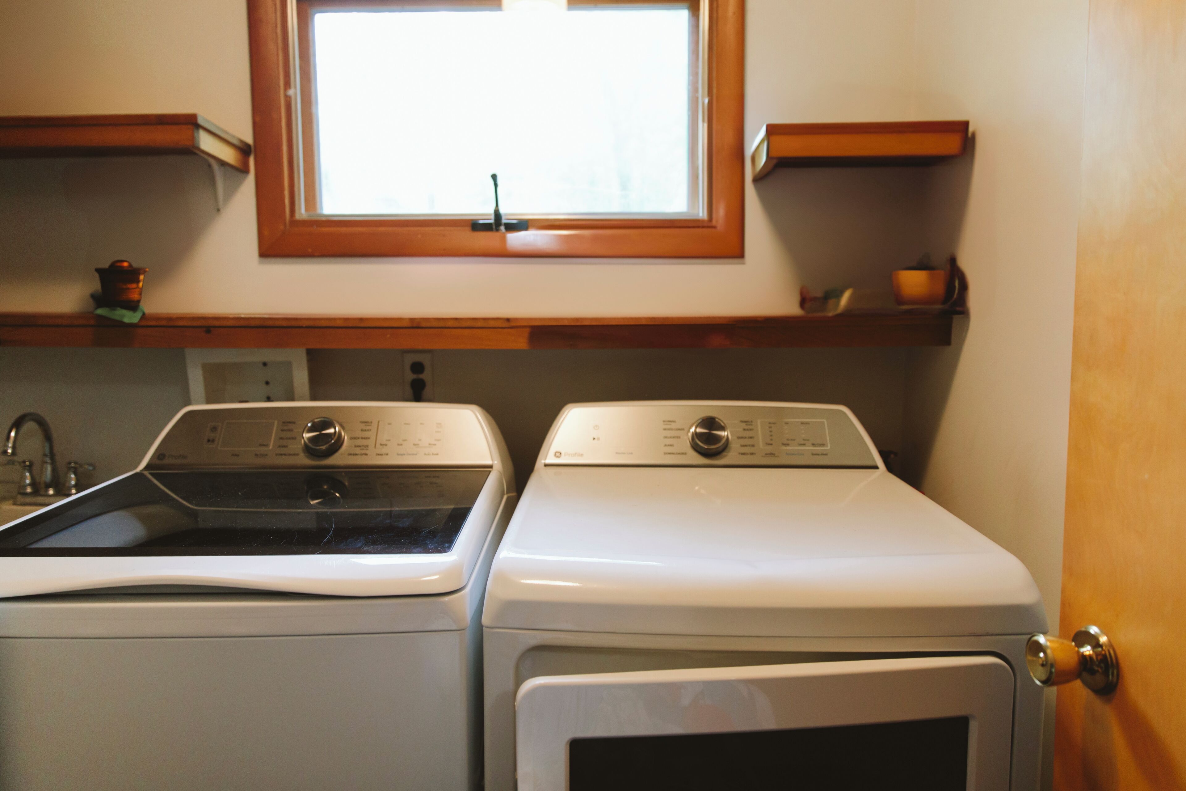 Un espace buanderie comprenant deux machines à laver, une fenêtre au-dessus et une étagère en bois sur laquelle se trouve une tasse.