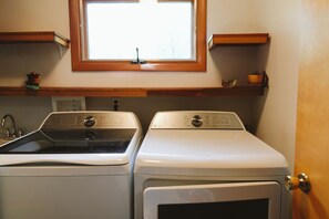 Un espace buanderie comprenant deux machines à laver, une fenêtre au-dessus et une étagère en bois sur laquelle se trouve une tasse.