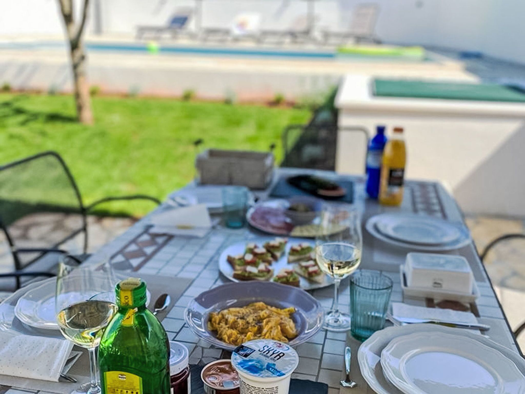 Et udendørs spisebord med forskellige retter, drikkevarer og en flaske olivenolie.