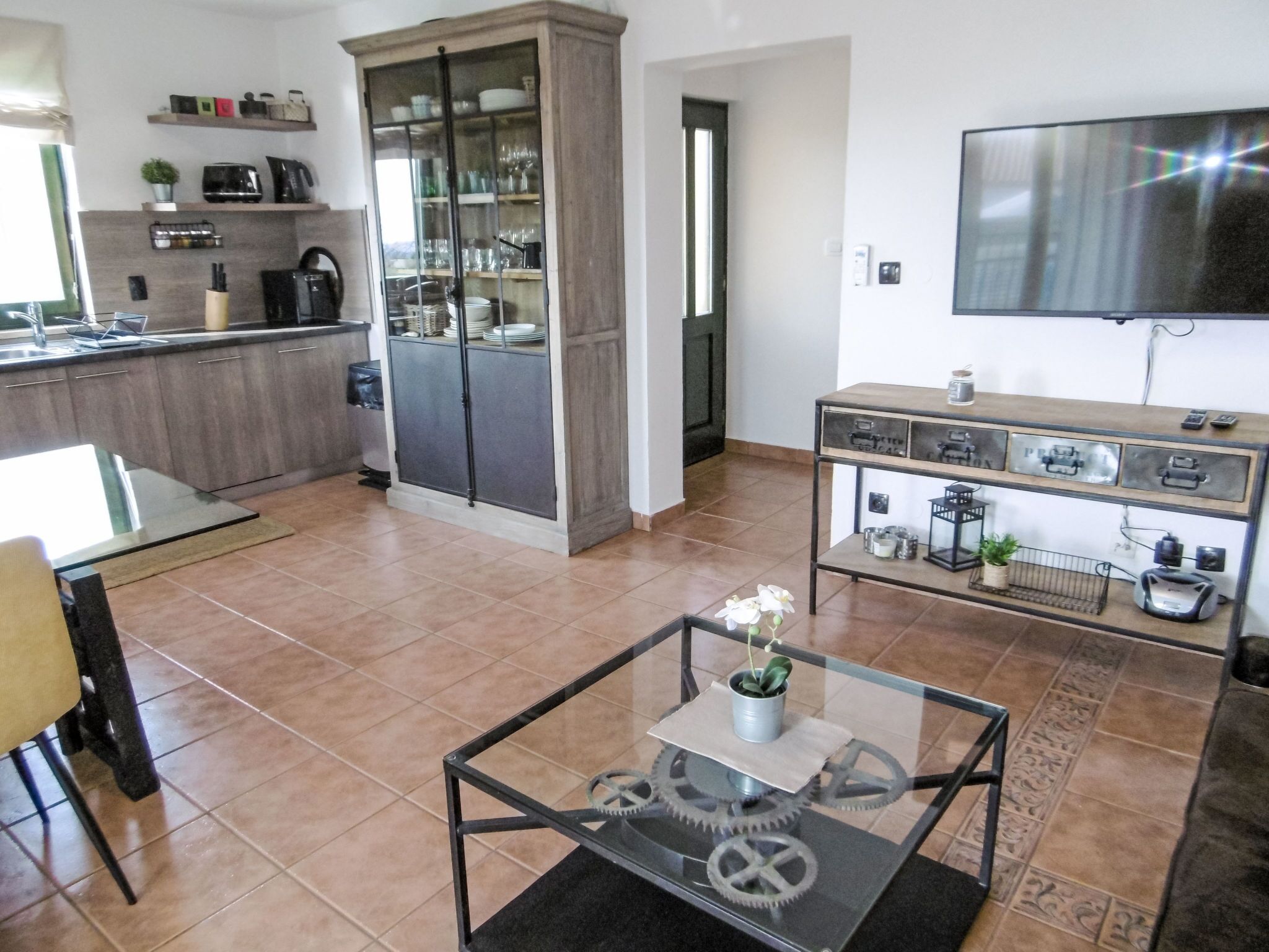 A modern kitchen with a built-in oven, a dining area with a glass table, and a TV mounted on the wall.