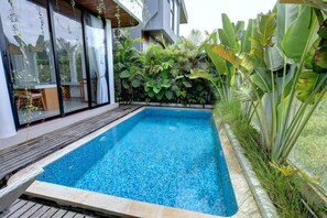 Una piscina rectangular con una terraza de madera alrededor, una puerta de cristal que da a una habitación y exuberante follaje tropical.