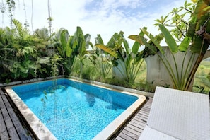 Piscina rectangular con agua cristalina azul, rodeada de exuberante vegetación tropical y una terraza de madera.