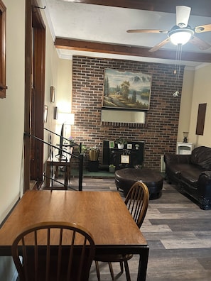 A dining area with a wooden table and chairs, a brick wall with a painting, a ceiling fan, and a sofa.