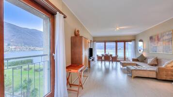 Een ruime woonkamer met een balkon met uitzicht op een meer en bergen, een houten kast, een televisie en een bank.