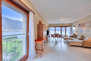 Een ruime woonkamer met een balkon met uitzicht op een meer en bergen, een houten kast, een televisie en een bank.