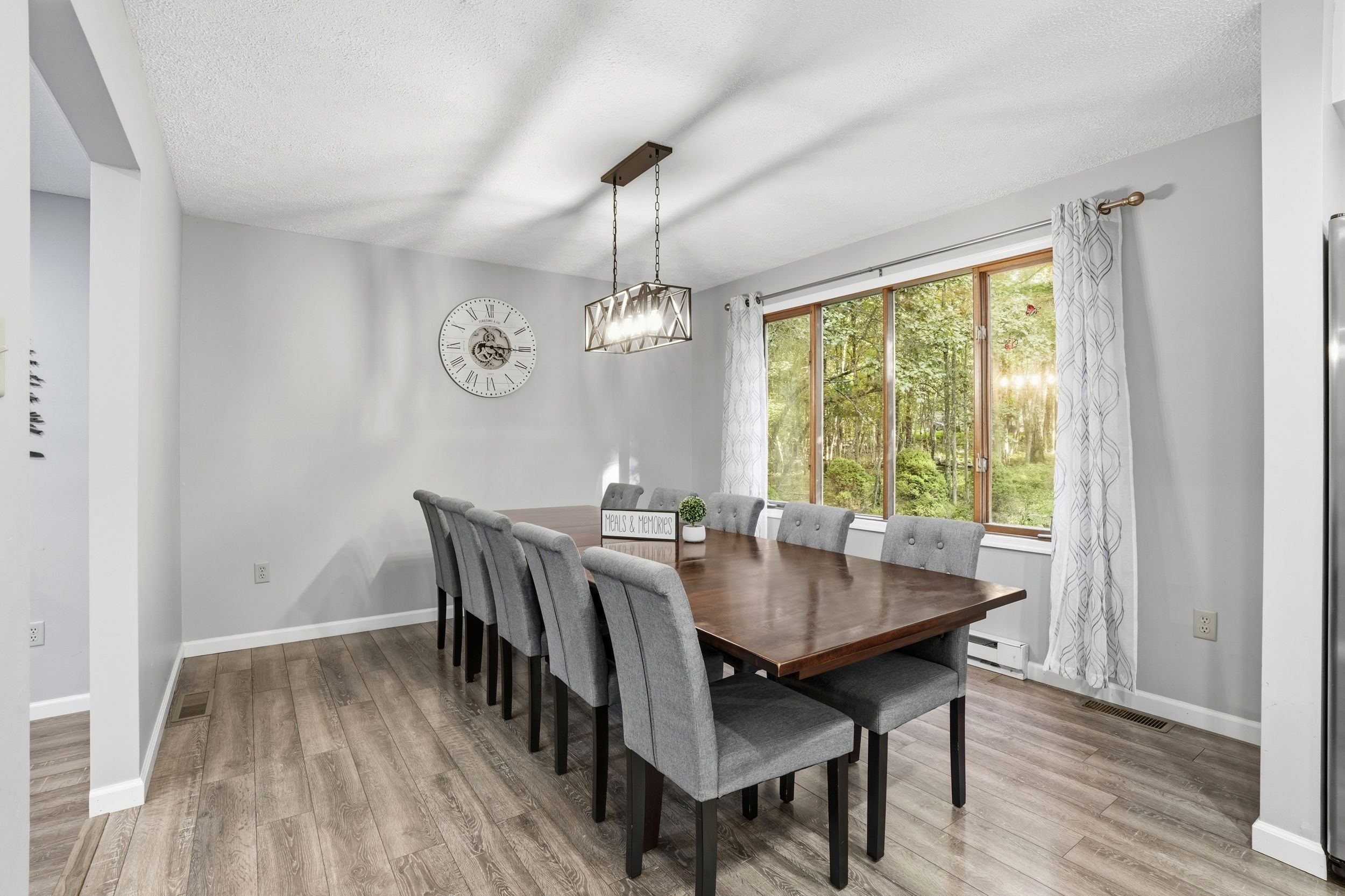 A dining room with a wooden table, grey upholstered chairs, a wall clock, and a large window with a view of trees.