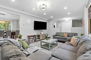 A modern living room with a flat-screen TV, grey sofas, a coffee table, and a dining area with a table and chairs. - The Birch: Poconos Getaway • Hot Tub, Game Room, Fire Pit • Sleeps 10 (Blakeslee)