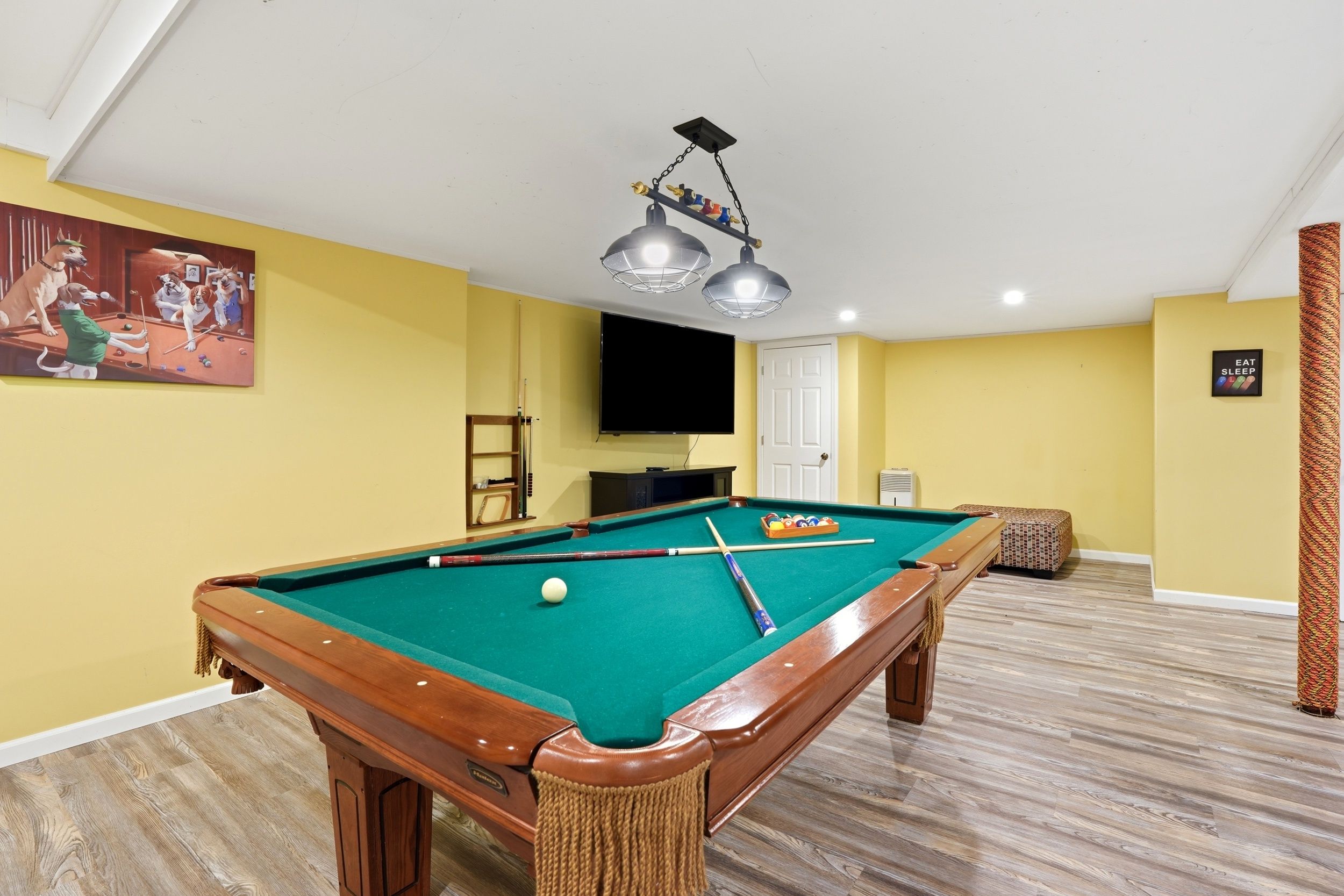 A pool table with a green felt surface, a television mounted on the wall, and a painting of dogs playing pool.